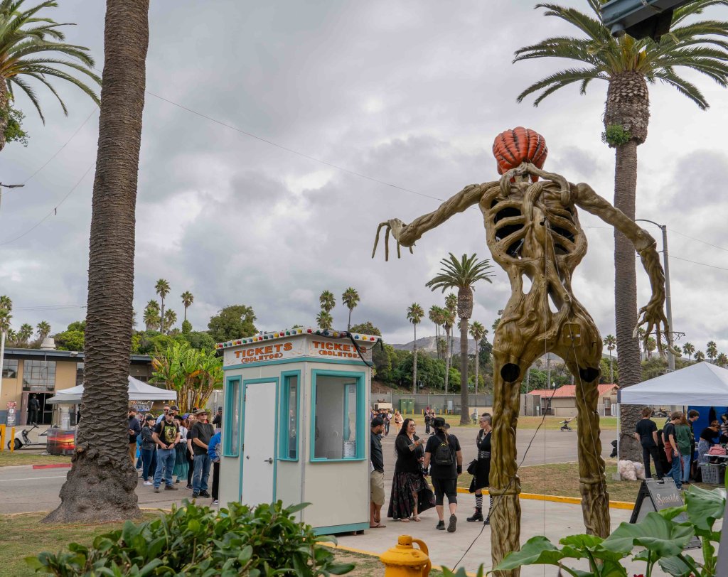 A skeletal pumpkin-headed sculpture stands near a ticket booth with people in line